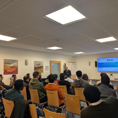 Eine Gruppe von Menschen sitzt in Stuhlreihen in einem Raum und schaut in die Richtung eines Screens  auf dem Zertifikatvergabe steht.