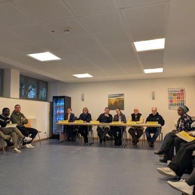 Eine Gruppe von Menschen sitzt im Stuhlkreis in U-Form. Vorne im Raum sind Tische aufgebaut hinter denen 6 weitere Personen sitzen.