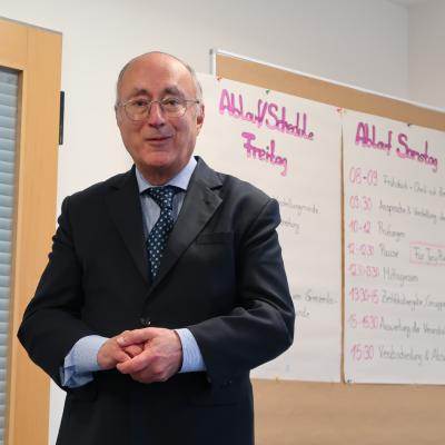 Ein Mann steht in einem Raum und schaut direkt in die Kamera, im Hintergrund sieht man auf einem Flipchart das Programm des Seminars. 