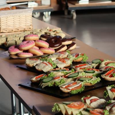 Tisch mit mehreren belegten Brötchen mit Salat, Gurkenscheiben, Tomaten und Käse sowie einem Stapel Donuts mit rosa Zuckerguss und Schokoladenglasur.