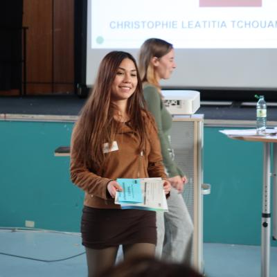 Junge Frau mit braunem Oberteil und schwarzem Rock hält mehrere blaue Broschüren in einem Veranstaltungsraum in der Hand. Im Hintergrund ist eine zweite Person zu sehen, die in die entgegengesetzte Richtung läuft.