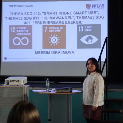 Eine Person steht vor einer Leinwand, auf der eine Präsentation gezeigt wird. Die Präsentation zeigt drei farbige Quadrate mit Symbolen und Texten zu den Themen nachhaltige Entwicklung. Vor der Leinwand steht ein runder Tisch mit einigen Gegenständen, darunter eine Wasserflasche.