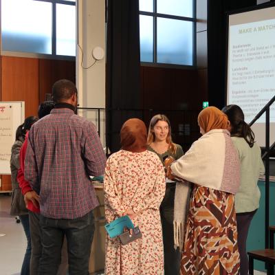 Eine Gruppe von Menschen steht in einem Kreis in einem Raum mit großen Fenstern und einer Holzverkleidung an der Wand. Einige Personen tragen Kopftücher, eine Person hält ein blaues Notizbuch hinter dem Rücken. Im Hintergrund ist eine weiße Tafel mit rotem Text und eine Projektionsfläche mit Text zu sehen.