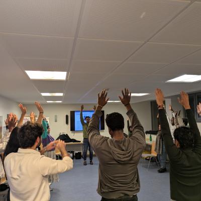 Eine Gruppe von Menschen steht im Kreis in einem Raum und alle heben ihre Hände in die Luft. 