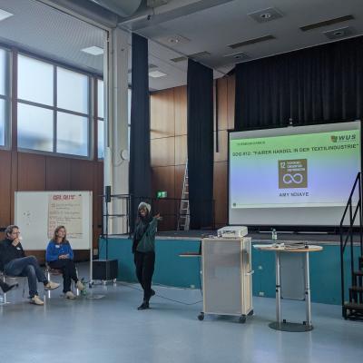 In einem großen Raum mit hohen Fenstern und Holzverkleidung an den Wänden steht eine Person vor einer Bühne und zeigt auf eine Leinwand, auf der eine Präsentation zu sehen ist. Vor der Bühne sitzen mehrere Personen auf Stühlen und hören zu.