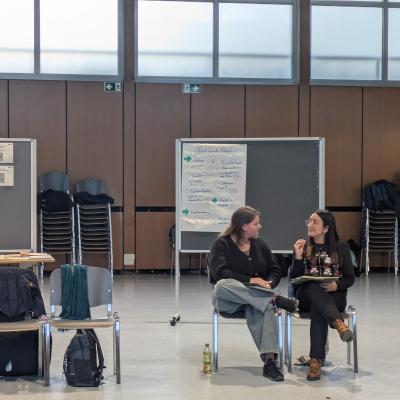 Zwei Personen sitzen auf Stühlen vor einem Flipchart in einem großen Raum mit Stühlen und Tischen und unterhalten sich.