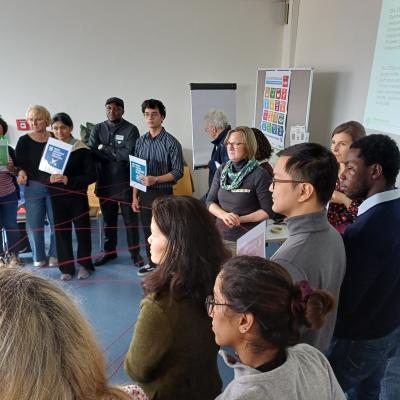 Eine Gruppe von Menschen steht im Kreis und hält bunte SDGs in der Hand und verbindet die SDGs durch ein Netz von Fäden