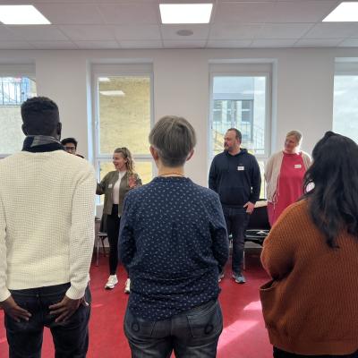 Eine Gruppe von Menschen steht in einem Raum. Im Vordergrund des Bildes sieht man die Rücken von zwei Menschen und im Hintergrund sind die Menschen der Kamera zugewandt. 