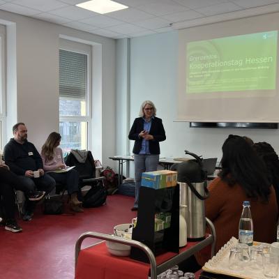 Eine Person steht vor einem Bildschirm und hält einen Vortrag. Auf dem Bildschirm steht Kooperationstag Hessen. 