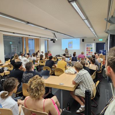 Zu Sehen ist ein Raum mit Studierenden, die einer tafel zugewandt sind. Voorne steht eine Person. 
