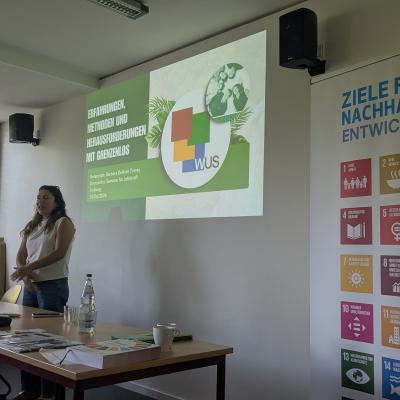 Eine Person steht vor einer Tafel und scheint zu reden. Rechts im Bild steht ein Plakat mit den SDGS. 