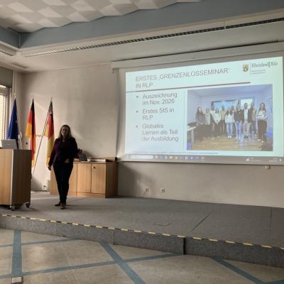 Die BNE Landeskoordinatorin aus Rheinland-Pfalz (Anna Bauer) steht auf einer Bühne. Auf ihrer Präsentation ist ein Foto zu sehen das eine Gruppe Menschen am Studienseminar Neuwied gemeinsam mit Grenzenlos-Referentinnen zeigt. 