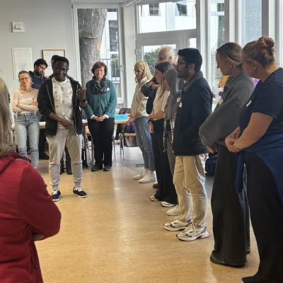Eine Gruppe von Menschen steht in einem Raum im Halbkreis. In der Mitte steht eine Person, die etwas zu sagen scheint.