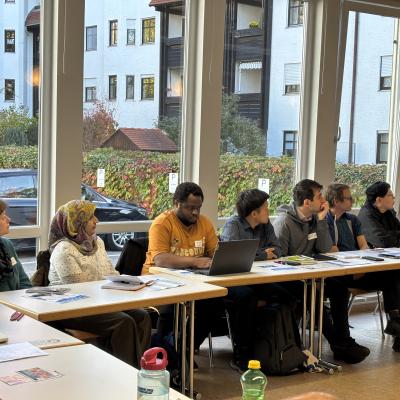 Zu Sehen ist ein Auschnitt aus einem Raum mit, wo einige Personen an einem Tisch sitzen und der vortragenden Person zuzuhören scheinen.