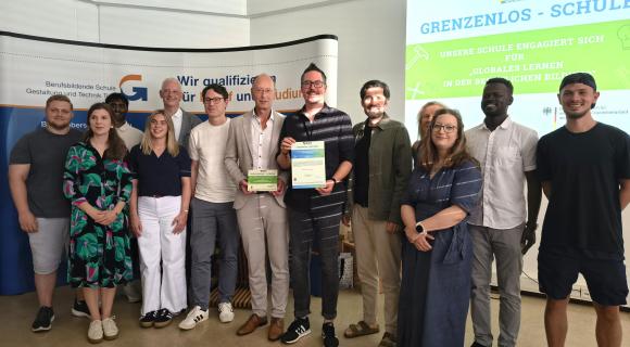 Eine Gruppe von Menschen steht vor einem Banner der Berufsbildenden Schule Gestaltung und Technik Trier. Der Schulleiter Stefan Justinger und Lehrkraft Michael Gent halten lächelnd die Grenzenlos-Plakette und Urkunde