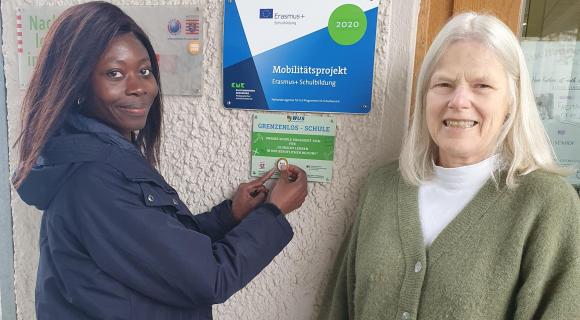 Zwei Frauen stehen an einer Hauswand mit mehreren Schildern, eine Frau befestigt einen Sticker auf einem grünes Schild mit Text.