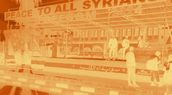 Foto einer Bühne mit dem Schriftzug „Peace to all Syrians – Freedom 2024“. Mehrere Menschen stehen oder gehen davor. Im Hintergrund Gebäude und eine Palme. Das gesamte Bild ist in orangefarbener Negativoptik gestaltet.