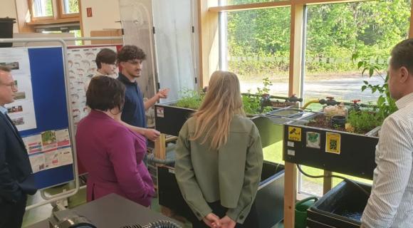In einem hellen Innenraum mit großen Fenstern präsentieren zwei Jugendliche einer Gruppe eine Aquaponik-Anlage mit Wasserbecken, Pflanzen und technischen Komponenten. Mehrere Erwachsene und Jugendliche betrachten die Anlage.