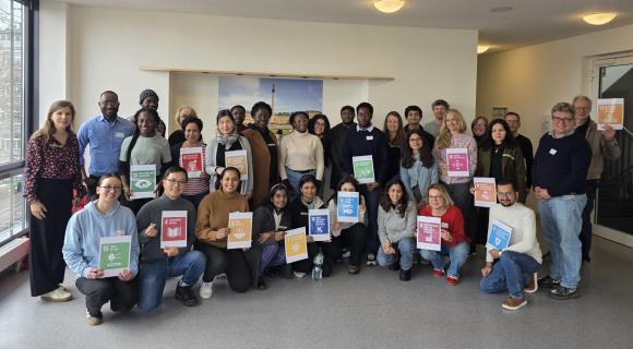 Eine Gruppe von Lehrkräften und internationalen Studierenden steht zusammen vor einer Wand sie halten die bunten SDGs hoch