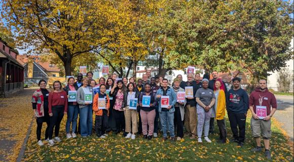 Eine Gruppe von 31 Menschen steht draußen vor bunten Bäumen. 