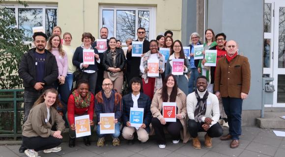 Eine Gruppe von Menschen stehen vor einem Haus und halten bunte Schilder mit den SDGs in den Händen