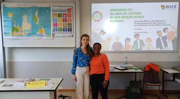 Zwei Frauen stehen vor einer Weltkarte und einer Leinwand, sie haben den Arm um sich gelegt. Links steht die Grenzenlos Referentin Helen Danso, rechts die Grenzenlos-Aktive Abla-Sika Hanou Gbekley. Auf der Leinwand ist das Grenzenlos Logo projeziert.