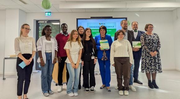Eine Gruppe von Menschen steht in der Aula der Schule. Einige halten Zertifizierungs-urkunden in der Hand.