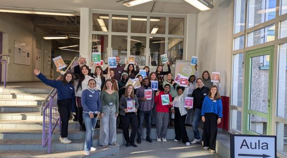 Eine Gruppe von 23 Personen, unterschiedlich in Alter, Geschlecht und Herkunft, steht auf und vor einem Treppenabsatz in einem hellen Innenraum. Viele lächeln in die Kamera und halten bunte Schilder mit Symbolen und Texten hoch, die die 17 Ziele für nachhaltige Entwicklung (SDGs) der Vereinten Nationen darstellen. Die Personen tragen lässige, unterschiedliche Kleidung, einige haben Namensschilder. Im Hintergrund ist ein Bürobereich mit Fenstern, Pflanzen und Informationsmaterial zu sehen. Die Stimmung wirkt