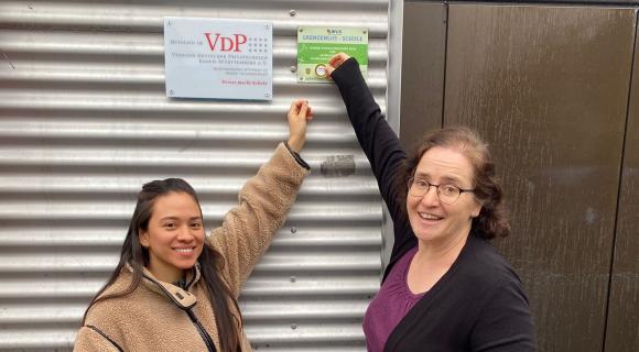 Zwei Personen stehen vor einer Metallwand mit drei Schildern. Eine Person zeigt auf ein grünes Schild.