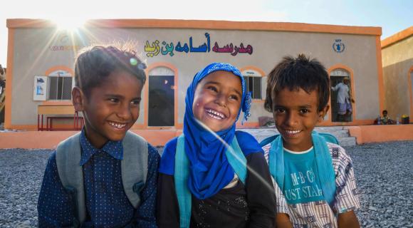 Drei lachende Kinder mit Schulranzen sitzen vor einem Gebäude mit arabischer Schrift und dem Logo des Welternährungsprogramms (WFP). Die Sonne scheint hinter dem Dach hervor. Das Bild vermittelt Freude und Bildungschancen.